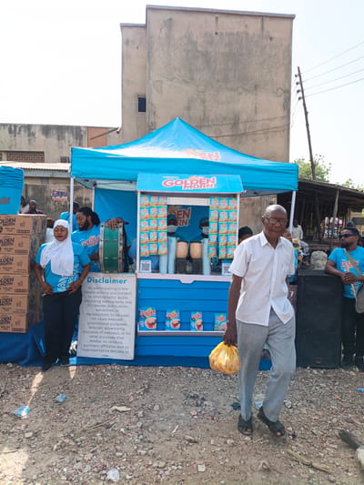 Here is a description of the image:
The image shows a "Golden Morn" promotional stand in what appears to be a street scene, likely in the city of Kawaji, Nigeria. The stand is predominantly blue and features the "Golden Morn" brand logo and cereal packaging.
Several people are visible. There is a group of people wearing matching "Golden Morn" t-shirts staffing the stand. A woman in a white headscarf is near the stand. An older man is walking in front of the stand, carrying a yellow bag.
The environment is somewhat cluttered. Some boxes of "Golden Morn" cereal are stacked next to the booth. The ground appears to be a dirt road. There are some simple buildings and an old power pole in the background. A large black speaker is near the booth, and a man in sunglasses is standing nearby with a drum.