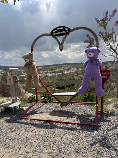 The image depicts a tourist attraction in Göreme, Turkey. A metal frame in the shape of a heart is adorned with two stuffed teddy bears, one purple and one tan. The frame has a sign in Turkish. The background shows a landscape of rock formations and valleys under a cloudy sky. A pay-per-view telescope is present near the base of the frame, positioned to view the surrounding landscape. The ground around the frame is covered with small pebbles.