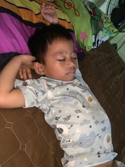A young child with dark hair is sleeping on their back, with their right arm bent and head resting on their right arm. They are wearing a light blue pajama set with a repeating pattern of cartoon elephants, clouds, and stars. The child is on a brown quilted surface, possibly a mattress or blanket. In the background, there are pillows with colorful striped and cartoonish patterns, and a black electric fan is visible in the upper right corner. The scene appears to be an indoor setting, likely a bedroom. The location is described as Sungai Meriam, Indonesia.