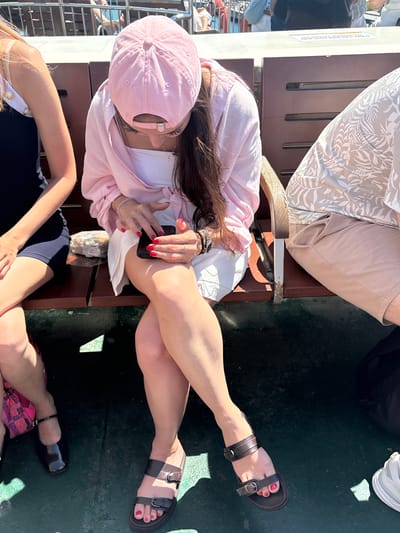 In Kadıköy, Turkey, three individuals are seated on a ferry. The person in the foreground, a woman with long dark hair, wears a pink baseball cap, a pink open button-up shirt over a white top, and a white skirt. She is looking down at her black smartphone, which she holds in her hands. Her toenails are painted red, and she wears dark brown sandals. To her left, another woman, partially visible, wears a dark blue sleeveless dress. She has a black patent leather flat shoe on her left foot. To the right of the main subject, a person in a light-colored tropical print shirt and light brown shorts is seated, mostly out of frame. The seating appears to be on a ferry, with wooden benches and a dark floor. Sunlight casts shadows on the floor. A wrapped food item rests on the bench between the two women on the left. The background shows railings and blurred figures of other passengers.
