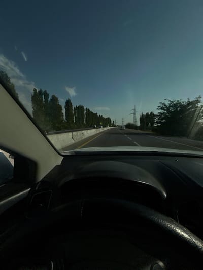 The image is a first-person perspective from inside a vehicle, looking out through the windshield and side window. The vehicle is traveling on a highway. In the foreground, the dark interior of the car, including the dashboard and steering wheel, is visible. The road ahead is paved, with white lane markings. To the left of the road, there is a concrete barrier, and beyond that, a line of tall, slender trees. To the right of the road, there are more trees and power lines with tall electrical towers. The sky is a clear blue with some wispy white clouds. The sun appears to be setting or rising, casting a bright light that creates some lens flare and highlights on the right side of the image. There are other vehicles visible on the highway in the distance. The overall impression is that of a scenic drive on a highway lined with trees and power infrastructure. The location is specified as the city of Istiqlol, Uzbekistan.