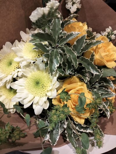 A bouquet of flowers is presented in a brown paper wrapping. The bouquet consists of white chrysanthemums with yellow centers, yellow roses, greenery with white-edged leaves, and some small white flowers. There is also greenery with small green buds. The image does not provide any information about a location or city.