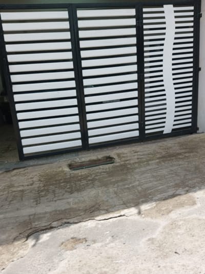 This image captures a black-framed gate with three panels, each filled with horizontal white slats, situated in Viengthong, Laos. The rightmost panel features an additional vertical, undulating white element overlaid on its slats. Directly in front of the gate, a rectangular, recessed drain opening with visible debris is present in the concrete pavement. The concrete ground exhibits a prominent linear crack extending across the lower foreground, alongside several darker, irregular patches indicative of moisture or staining. No individuals, actions, or interactions are visible.