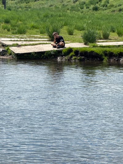 A young boy in a black t-shirt and shorts is sitting on a concrete platform next to a body of water. He is resting his chin on his hand and looking down. The platform is bordered by green grass and some yellow wildflowers. The water is rippling and reflects the blue sky. In the background, there is more green grass and some bushy plants. The scene appears to be in an outdoor recreational area.