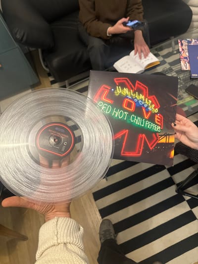 A vinyl record and its cover are displayed. The record is a clear translucent pressing of Red Hot Chili Peppers' "Unlimited Love" album. The album cover features a neon sign design with the album's title and band name. The record is held in a hand, while the album cover rests on a surface. A person with a phone is also in view, along with parts of a black and white striped rug and furniture.