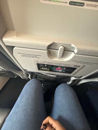 The image shows an airplane seat with a tray table in the upright position. The tray table has instructions and advertisements, including a Hertz advertisement offering car rentals for $100 per day. Part of a passenger wearing blue jeans is also visible. The image does not contain information to place it in San Antonio, United States of America.