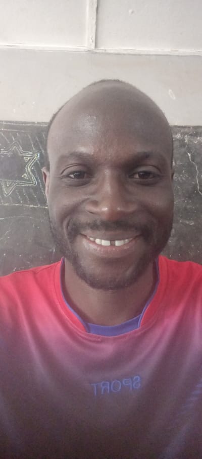 An adult male subject is depicted in a close-up, head-and-shoulders portrait, positioned indoors in Barnawa, Nigeria. The individual has a dark complexion, a receding hairline, and maintains a short beard and mustache. He is smiling directly at the camera, displaying a visible diastema between his two upper front incisors. He wears a short-sleeved garment, primarily reddish-pink/coral, with a blue/purple contrasting collar visible underneath. The word "SPORT" is legible in reverse text on the lower front portion of his shirt. The background to the left of the subject features a dark surface with a white chalk outline of a Star of David. To the right, the background consists of a lighter, possibly textured wall. The upper background shows a white ceiling or upper wall section with visible structural lines.