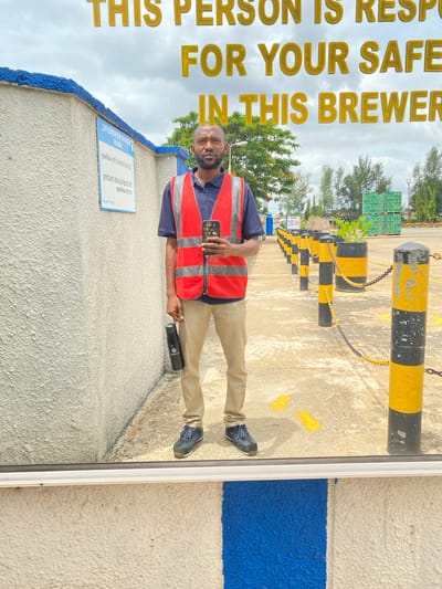 The image shows a man wearing a dark blue polo shirt and a bright orange safety vest with reflective stripes. He has a beard and is smiling. He is holding a black water bottle in his right hand and a smartphone in his left. The man is standing outdoors on a paved surface. In the background, there are rows of black and yellow bollards with chains, and stacks of green crates, possibly containing beverage products. There are also trees and a cloudy sky. A sign is visible on a textured, light-colored wall with a blue stripe. The sign has text in both English and another language, but the text is difficult to read due to the angle and resolution. Large, golden, block letters on the left side of the image read "THIS PERSON IS RESP FOR YOUR SAFE IN THIS BREWER". The overall scene suggests an industrial or operational setting, likely a brewery or distribution center. The man appears approachable and content. The lighting suggests it is daytime.