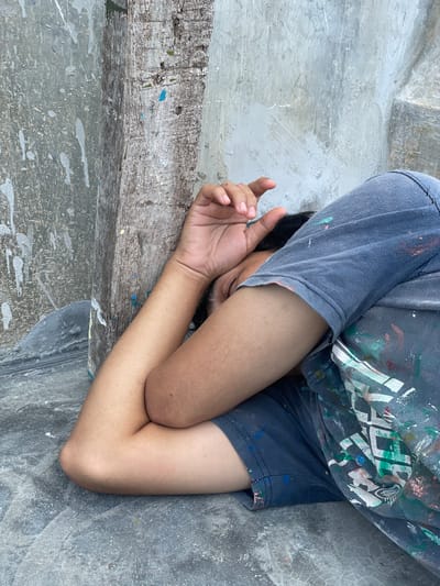 An individual lies on their right side on a dark grey concrete floor, resting against a weathered, vertically oriented wooden post and a light grey unfinished concrete wall. The person's face is obscured, turned away from the camera and partially covered by their right hand. They wear a dark grey short-sleeved t-shirt and dark blue shorts, both heavily stained with various colors of paint (e.g., blue, green, white, pink). The wooden post also shows blue paint splatters. The concrete wall in the background has a textured, unpainted finish with subtle paint marks, and a stepped concrete element is visible in the upper right. The environment appears to be a utilitarian or construction-related space. The specific location within Mns Asan, Indonesia, cannot be determined from the visual information provided.