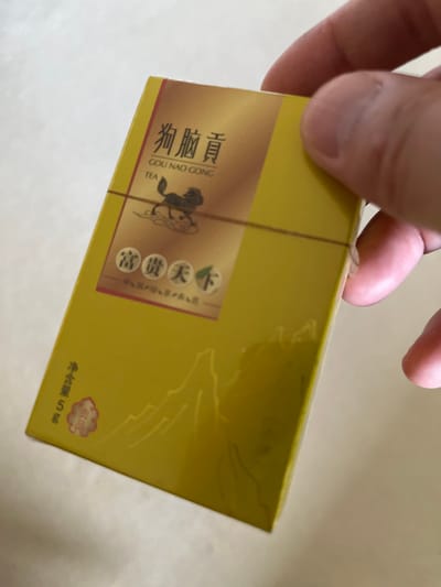 A close-up, slightly angled shot captures a hand holding a rectangular, golden-yellow box, appearing to be sealed in clear plastic. The background is a plain, light-colored surface, suggesting an indoor setting. The box features a prominent brown vertical stripe on its left side, where most of the visual information is concentrated.

Within this brown stripe, Chinese characters and Pinyin romanization are clearly visible. The main text reads "狗脑贡" (Gou Nao Gong) with its Pinyin transcription "COU NAO CONG" directly below, followed by the English word "TEA", indicating the product's nature. A stylized graphic of a dark, possibly mythical animal, reminiscent of a horse, is positioned next to this text. Further text on the box includes "富贵天下" (Fugui Tianxia), which translates to "Wealth and Honor Under Heaven", and "中国红" (Zhongguo Hong), meaning "China Red". A small red, seal-like stamp on the lower right of the box contains the characters "贡品" (Gong Pin), signifying a "Tribute Product". Below this, the net weight "净含量 5g" (Jing Hanliang 5g) is printed. The clear plastic wrapping shows some minor creases, indicating the product is new and unopened. The lighting is bright and even, highlighting the details of the product's packaging.