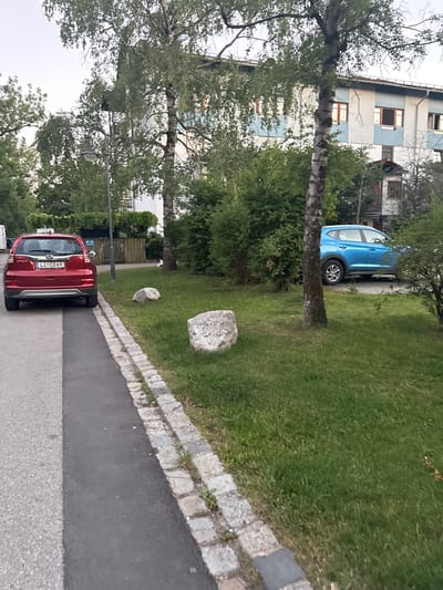 In the city of Höhenkirchen-Siegertsbrunn, Germany, a red SUV is parked on the left side of a paved path. The vehicle's license plate reads "LI CD69". A blue hatchback is parked to the right of the path, slightly further down. The scene is a residential area with a building in the background, surrounded by trees and green grass. Two large rocks are visible on the grass. The sky is overcast, suggesting it might be evening or a cloudy day.
