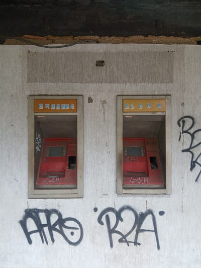 The image depicts two abandoned ATM machines in Guayana City, Venezuela. The ATMs are red and appear to be older models, with some graffiti visible on their surfaces and the surrounding wall. The building facade is made of textured concrete, and above the ATMs, there is a dark, rough ceiling or overhang. The scene suggests a state of disrepair and neglect, possibly in an outdoor or semi-outdoor urban setting. There are no people visible in the image. The overall impression is one of abandonment and decay.
