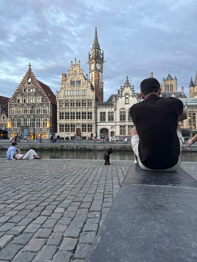 The image depicts a scenic view of the Graslei and Korenlei canals in Ghent, Belgium, under a cloudy sky. In the foreground, a person is sitting on a stone bench, facing away from the camera, with a small brown dog on a leash in front of them. The dog is looking towards the canal. Further to the left, another person is sitting on the cobblestone ground, leaning back and looking towards the canal. The iconic historical buildings of Ghent, characterized by their gabled facades and ornate architecture, line the canal's edge. Many windows of these buildings are illuminated, suggesting the time is late afternoon or early evening. A few other individuals are visible walking or sitting along the canal promenade. The cobblestone pavement in the foreground is well-maintained, leading towards the water's edge.