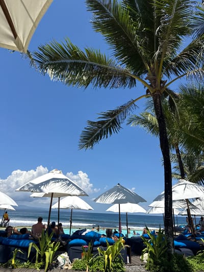 The image shows a beach scene, possibly a resort, in Medahan, Indonesia. The camera is angled upwards, capturing a view of the sky, palm trees, and beach umbrellas. The sky is blue with some white clouds. Several white beach umbrellas are visible, providing shade for people lounging in blue chairs. The ocean is in the background, with people swimming. Lush greenery is present on the right side of the frame. People are lounging on the chairs and in the water.