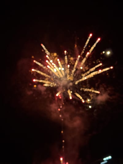 This forensic image captures a nocturnal scene in Debrashtitsa, Bulgaria, depicting a fireworks display against a dark sky. The central focus is a luminous burst of golden-yellow and red light trails, extending radially outward, characteristic of an exploding pyrotechnic device. The light sources exhibit a distinct bokeh effect, appearing as blurred circular or oval shapes due to the shallow depth of field. Reddish-brown smoke and haze are visible within the dark background, particularly around the core of the fireworks burst and trailing downwards. A singular, faint, glowing stream extends from the lower portion of the primary explosion towards the bottom of the frame. The environment is uniformly dark, consistent with nighttime conditions, with no discernible people, structures, or specific landscape features. A few extremely blurred, distant artificial lights are faintly visible in the far background, primarily in the upper right quadrant.