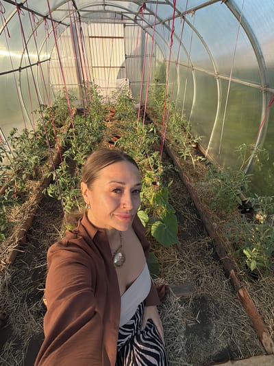 The image shows a woman in a greenhouse filled with plants. The greenhouse appears to be a tunnel-style structure with clear plastic sheeting. The ground inside is covered with straw or hay. There are rows of plants, likely tomato plants, with red strings tied to them for support. The woman is standing in the foreground, taking a selfie. She is wearing a brown shirt and a white top, with a patterned skirt. She has jewelry on, including a necklace and earrings. The sunlight is coming in from the side, creating a warm glow. The background shows the interior of the greenhouse, with more plants and the structure's framework. There is a door or entrance at the far end of the greenhouse. There is no visible indication that this location is in Kansk, Russia.