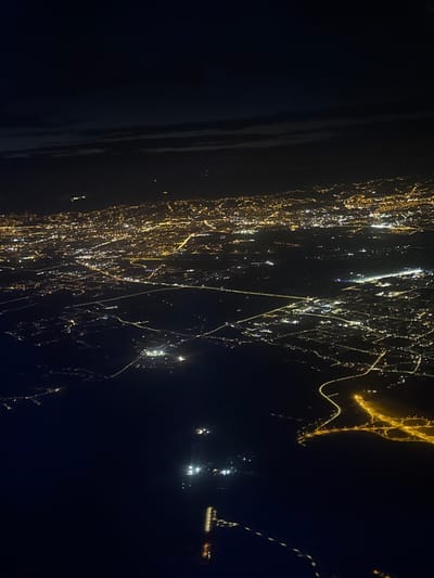 This is an aerial night view of Surčin Urban Municipality, Serbia. The image captures a vast expanse of urban lights spread across the landscape, indicating a densely populated area. The lights, primarily yellow and white, illuminate the city's layout, roads, and possibly buildings from a high altitude. The darkness of the night sky contrasts sharply with the illuminated urban sprawl. The perspective suggests the photo was taken from an airplane or a similar elevated vantage point. The general environment is a city at night, with no discernible natural elements like mountains or large bodies of water clearly visible, other than the dark expanse which may be a river or a large area of low-lying land. The scene is devoid of any specific human presence, vehicles, or individual structures that can be clearly identified from this altitude and lighting. The overall impression is that of an extensive, illuminated urban network.