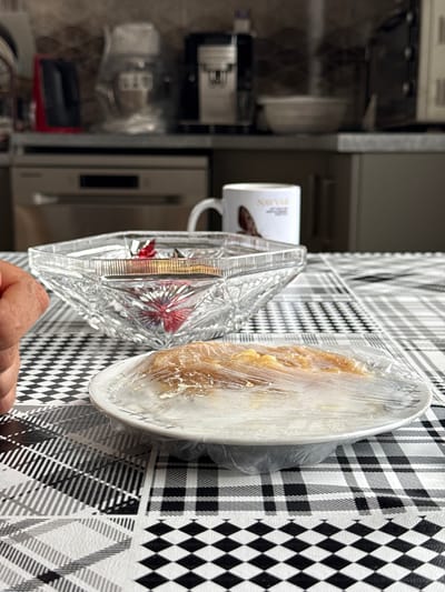 The image shows a table setting in what appears to be a kitchen or dining area. On the table, which is covered with a black and white checkered tablecloth, there is a clear crystal bowl filled with some sort of wrapped candies or chocolates. Next to it, there is a white mug with a picture of a coffee bean and text on it. In front of these items, a white plate holds a dessert, possibly a pastry or cake, covered in plastic wrap. The background suggests a kitchen environment with appliances like a coffee machine and a dishwasher visible. There is a person's hand visible on the left side of the frame, reaching towards the table. The overall impression is a casual meal or snack setting. The location in Khazar Raion, Azerbaijan, is not visually discernible from the image itself.