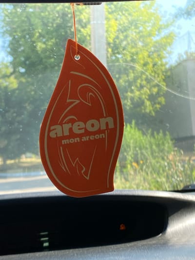 An orange teardrop-shaped air freshener with the brand name "areon" in white text and a stylized design is hanging from a string in front of a car windshield. The background visible through the windshield is blurred, showing green trees and foliage, suggesting an outdoor setting. The air freshener is positioned slightly off-center to the left of the image. Below the windshield, a portion of a car's dashboard is visible, with some indicator lights illuminated. The overall scene appears to be captured from inside a vehicle. There are no people or specific landmarks identifiable that would definitively place the location in Bender, Moldova.