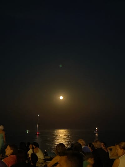 The image depicts a nighttime scene on the waterfront of Canet de Mar, Spain. A large, bright moon is visible in the sky, casting a shimmering reflection on the calm sea. Several boats are on the water, their lights twinkling in the distance. In the foreground, a group of people are gathered on the shore, sitting and standing, many facing the water and the moon. Their faces are partially illuminated by the moonlight and possibly artificial lights from the gathering or surrounding areas. The atmosphere appears relaxed and social, with individuals engaged in conversation or simply enjoying the view. The dark sky and the illuminated water create a serene and somewhat magical ambiance.