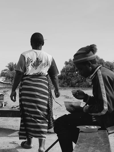 The image captures an outdoor scene in Kirana, Nigeria, under a bright, clear sky. In the foreground on the right, an individual is seated on a low, dark wooden bench, observed in profile facing left. This person wears a dark long-sleeved jacket with light stripes on the sleeves and a dark beanie hat with a tassel. They hold a small, light-colored bowl to their mouth with their right hand, consuming its contents, while a larger, light-colored bowl rests in their left hand on their lap. A third, flat, light-colored plate holding several spherical food items is positioned on the bench beside them. In the mid-ground on the left, a second individual is seen from behind, walking away from the camera. This person wears a light-colored t-shirt with multiple dark graphic designs on the back, resembling wrapped candies, each bearing the text "TOM TOM." Below the t-shirt, a dark, striped wrapper or skirt extends to their ankles, and they wear light-colored open-toed footwear. The background features dense green foliage from trees and a faint outline of a structure or fence, with the ground appearing unpaved.