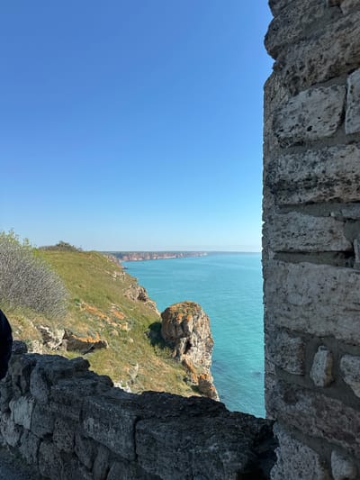 The image shows a view from a stone structure in Balgarevo, Bulgaria. The frame is formed by stone walls at the top and right side of the image. The view looks out over the Black Sea, with a coastline visible in the distance. A rocky cliffside covered with grass and vegetation is seen on the right, sloping towards the sea. The sky is clear blue.