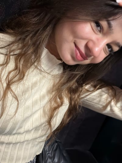 A young woman with long, wavy brown hair smiles softly at the camera. She is wearing a textured cream-colored sweater. She rests against a dark surface, possibly a couch or chair. Her skin is smooth and her lips are a soft pink color. Her expression is relaxed and inviting. The light catches her face, highlighting her cheekbones and the slight curve of her nose.