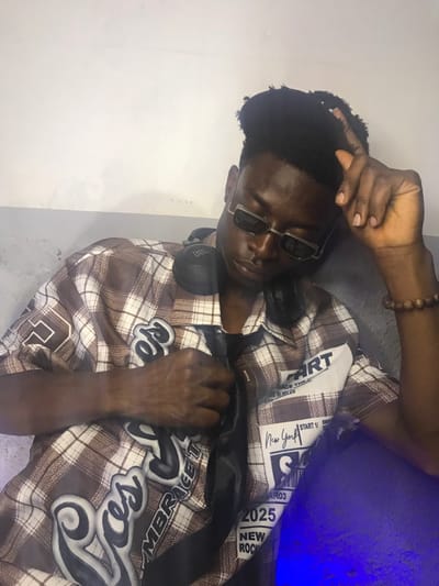 A young Black male is depicted in an indoor setting in Kirana, Nigeria, positioned against a light-colored wall with a darker gray lower section. He wears a short-sleeved brown and white plaid button-up shirt featuring multiple stitched patches, including "ROSES," "EMBRACE," "START," "New York," "2025," and "NEW ROCK." A black satin tie is visible beneath his shirt collar. He wears rectangular dark sunglasses and black JBL over-ear headphones resting around his neck. A brown beaded bracelet is on his right wrist. His dark hair is styled with volume on top. His head is tilted slightly to the right, and his eyes are closed or directed downward. His left hand is raised, fingers slightly parted, near the left side of his head. His right hand is positioned on his chest, lightly gripping a section of his shirt and tie. A distinct blue light source illuminates the lower right portion of the frame, casting a blue glow on his shirt.