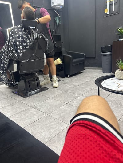 The image shows a barber shop in Petrich, Bulgaria. A person is sitting in a barber chair while another person, presumably the barber, is cutting their hair. The barber is wearing a purple t-shirt and a dark apron. The person in the chair is wearing a black cape with a white pattern of scissors and comb icons. The shop has black walls, a tiled floor, and modern furniture, including a black leather armchair and a coffee table. There is a trash can and a small potted plant in the background. The foreground shows the legs of another person wearing red basketball shorts, suggesting they are waiting their turn or observing.