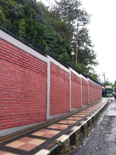 A long, segmented wall constructed from red-orange bricks with vertical grey concrete pillars and a grey top coping extends diagonally from the lower left towards the upper right, forming the left boundary of a street in Cipanas, Indonesia. Black razor wire is affixed along the entire top edge of the wall. Dense green foliage and trees are visible behind and above the wall, suggesting an elevated slope. A checkered sidewalk, comprised of alternating light brown and dark brown/red square tiles, runs parallel to the wall, slightly elevated from the adjacent wet asphalt road. Several wooden utility poles with multiple overhead wires are present against an overcast sky, extending down the street. In the distance, on the right side of the road, indistinct structures and a portion of a green vehicle are visible. No individuals are present.