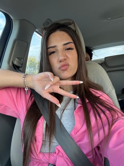 This image captures two individuals inside the interior of a vehicle during daylight hours. The primary subject is a female, positioned in the front passenger seat, actively taking a selfie. She has long, dark brown hair and fair skin, with visible makeup including enhanced eyelashes and defined eyebrows. Her lips are puckered in an expressive pose, and sunglasses rest on her head. She is attired in a light gray ribbed undergarment beneath a bright pink, long-sleeved button-up shirt. A dark seatbelt is securely fastened across her torso. On her left wrist, she wears a silver bracelet featuring small beads and charms. Her left hand is raised, displaying a "peace sign" (V-sign) gesture with extended index and middle fingers, revealing long, white-tipped fingernails. A second individual is partially visible in the rear seat, with only the top of their head and dark, curly hair showing above the front passenger seat's headrest. The car's interior components, including the headliner, side pillar, and seats, are light grey or beige. Through the vehicle's windows, a clear blue sky with sparse clouds and green tree foliage is observable. The image does not contain any discernible features or landmarks to confirm the specific location as Tsaratsovo, Bulgaria.