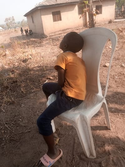 In Gudige Sabo, Nigeria, a young child, dark-skinned, is seated on a white plastic chair, viewed from the back-left side. The child wears an orange short-sleeved t-shirt, dark blue long shorts or capri pants, and light pink Adidas slide sandals. The child's head is tilted upward and to the left, looking into the distance.

The scene is an outdoor, dry, unpaved area characterized by reddish-brown soil and sparse dried vegetation. In the mid-ground, a single-story structure with light brown walls and a corrugated metal roof stands, featuring two rectangular windows with dark grates. Further in the background, to the left of the building, a group of approximately three to four smaller, indistinct figures, presumably children, are visible on the dry ground, appearing to be gathered or playing. The sky is clear and bright, indicating daytime.