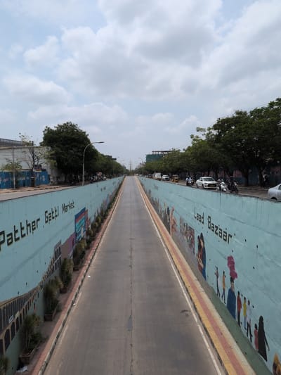 The image shows a view from above looking down a street lined with a turquoise retaining wall. The wall is decorated with murals of people and text that reads "Patther Gatti Market" on the left and "Laad Bazaar" on the right. The street itself is asphalt and appears to be a multi-lane road. There are cars, motorcycles, and auto-rickshaws driving on the road. Trees line the top of the retaining wall on both sides of the road, and the sky is partly cloudy. The scene appears to be in an urban environment, possibly a market area in Hyderabad, India, given the text on the wall which refers to well-known markets in Hyderabad.