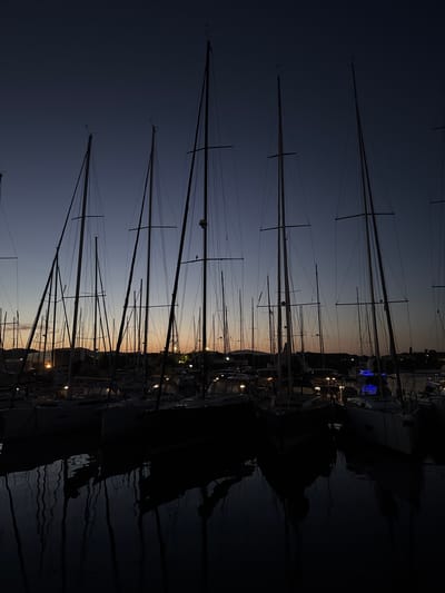 The image captures a marina at twilight, likely dusk, within the Lavreotiki Municipal Unit, Greece. Numerous sailboats are moored in still water, their tall masts and intricate rigging forming prominent dark silhouettes against the sky. The sky displays a gradient, transitioning from a deep navy blue at the top to a lighter blue-grey mid-frame, and finally to a warm band of orange, pink, and yellow hues along the horizon. The hulls of the sailboats are largely obscured by shadow, though various artificial lights, predominantly warm white or yellow, are visible on their decks and cabins. A distinct blue light is visible within the water on the right side of the frame. The calm water in the foreground reflects the vertical masts and some of the boat lights, creating elongated, shimmering distortions. In the background, a distant shoreline is visible with the dark silhouettes of what appear to be buildings or structures, punctuated by scattered small, glowing artificial lights. No human figures are present or discernible in the scene.