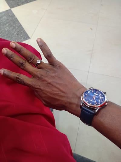 The image displays a close-up, high-angle view of a dark-skinned individual's left hand and forearm. A silver-colored ring with a prominent clear gemstone is worn on the fourth finger of the hand. On the left wrist, a watch with a rose gold-toned casing, a dark blue dial featuring multiple sub-dials and luminous markers, and a dark blue leather strap is visible. The individual wears a red garment, possibly a shirt or dress, exhibiting a subtle vertical striped texture. A sliver of a gray garment, likely trousers, is also discernible. The background consists of an indoor tiled floor, primarily light beige, with a section of dark gray and light beige checkerboard pattern in the upper left. The specific location within the city of Dorayi, Nigeria, cannot be determined from the visual information provided.
