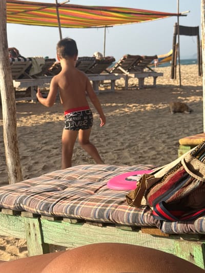The scene depicts a sandy beach in Bhagatwadi, India, under clear daytime conditions. In the immediate foreground, the perspective is from a reclining position on a wooden beach bed, displaying a plaid-patterned cushion, a pink frisbee, and a striped woven bag with a dark strap. A young child, appearing to be male, is observed from behind in the midground, walking leftward across the sand. He is shirtless, wearing black swim trunks with a red waistband and partially visible white text including "BEST SINCE 1997 BY G LIVE." His right arm is slightly elevated. In the background, several wooden sun loungers with cushions are positioned under a multi-colored striped canopy (red, yellow, green, blue). Another vertical wooden structure stands to the right. A small, dark-furred animal rests on the sand to the right of the child. The ocean is visible in the far background, with a yellow and blue boat partially present on the sand near the water's edge.