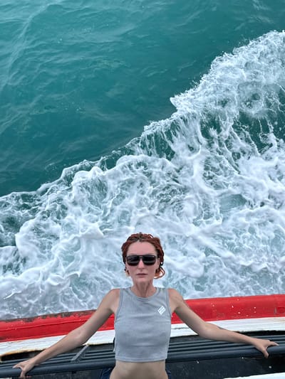 The media file shows a person with reddish-brown braided hair and sunglasses, wearing a grey crop top, leaning against a railing on a boat. They are looking towards the camera with a neutral expression. The person's right arm is extended upwards, holding onto the railing, while their left arm is bent and resting on their leg.
The scene is set on the water, with choppy, turquoise-colored waves and white foam creating a dynamic background. The boat has a red and white stripe along its edge, and a dark metal railing. The image appears to be taken from a high angle, looking down at the person and the water.
The weather seems clear and sunny, suggesting it is daytime. The vibrant colors of the water and the foam indicate movement and a potentially breezy atmosphere. The location cues, such as the tropical-looking water, suggest a warm climate.
There is a small white tag on the person's crop top with some faint text that is illegible in the image. The overall mood is relaxed and adventurous, with the person enjoying a boat trip.