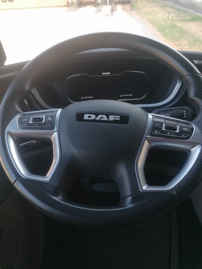 The image shows the interior of a truck cab, focusing on the steering wheel. The steering wheel is black with silver accents and features several buttons for cruise control, speed limiter, and audio controls. The DAF logo is prominently displayed on the center of the wheel. In the background, a digital dashboard displays the time as 10:05. The exterior environment is visible through the windshield, showing a sunlit outdoor scene with a paved surface and some greenery. The truck appears to be parked, possibly in an industrial or roadside area. The lighting suggests it is daytime. There are no people visible in the image. The image does not provide any specific information about the location being Santo Stefano Ticino, Italy.