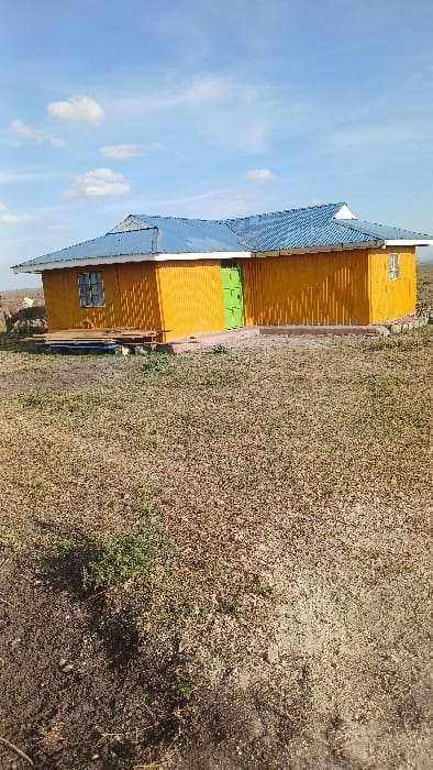 A single-story, rectangular building with bright orange vertical siding and a blue corrugated metal hip roof with white eaves trim is centrally positioned on dry, sparsely vegetated ground in Kaputiei North ward, Kenya. A lime green door is centrally located on the building's front facade. To the left of the green door, a window with a grid pattern is visible; another window is present to the right of the door. The building's foundation is a reddish-pink base. A small, low collection of wooden planks and other materials rests on the ground immediately to the left of the main entrance. The foreground and middle ground are composed of light brown soil interspersed with patches of dry green and yellow vegetation. The background features a flat horizon line under a clear blue sky with scattered white cumulus clouds. No individuals or animals are present in the visible scene.