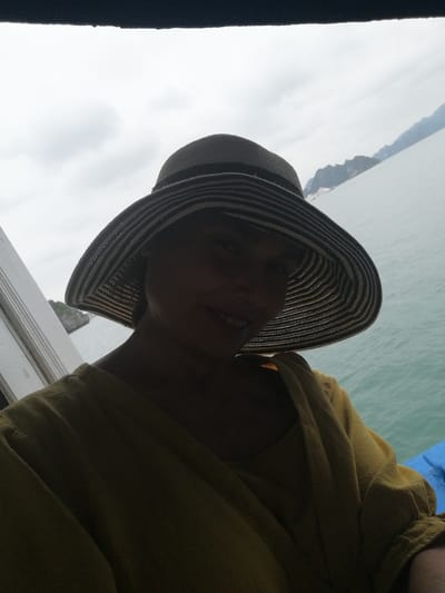 The image shows a person, likely female, photographed from a low angle, mostly obscured by shadow. She is wearing a wide-brimmed hat with alternating dark and lighter horizontal stripes and a mustard-yellow long-sleeved top or dress. A subtle smile is visible on the shadowed part of her face.

The subject is on a boat, indicated by a dark overhead structure at the top of the frame, white wooden or painted railings on the left, and a blue element, possibly a tarp or cushion, on the bottom right. The background features a large body of water, appearing calm with a light blue-green hue. In the distance, numerous distinct karst limestone islands or mountains rise dramatically from the water, partially obscured by a light atmospheric haze.

The sky is overcast and bright, suggesting daytime, but the direct sunlight is diffused, contributing to the deep shadows on the subject's face. The overall activity appears to be a leisurely boat ride or tour through a scenic bay. The prominent geological formations strongly suggest a location characteristic of Southeast Asia, such as Ha Long Bay. No text is visible in the image.