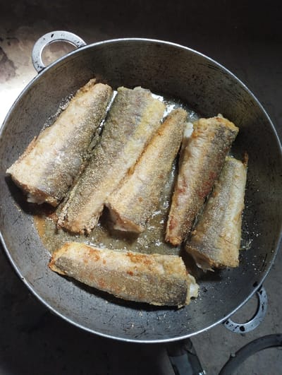 A close-up, overhead shot shows several pieces of breaded fish being fried in a dark, well-worn metal pot. The fish are golden brown and appear to be coated in a coarse meal or breadcrumbs. The pot is filled with oil, and some of the fish are partially submerged. The background is dark and indistinct, with a rough, textured surface visible at the top and bottom of the frame, suggesting an outdoor or rustic setting. The scene is likely in Fălești, Moldova, given the context provided, and depicts a simple, home-cooked meal in preparation.