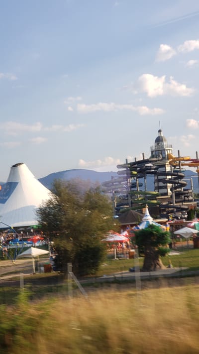 The image shows a water park in Bešeňová, Slovakia, with a large conical white tent structure on the left and a tall, multi-story water slide tower with multiple colorful tubes on the right. In the background, rolling hills are visible under a blue sky with scattered clouds. The foreground is blurred, showing dry grass and what appear to be fence posts. Several people are visible in the mid-ground, near the tent and the water slides, suggesting the park is open. Red and white striped awnings are present, along with some green foliage and trees. The overall impression is of a sunny day at a recreational facility.