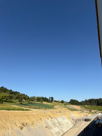 The image depicts a construction site in Skrzydlna, Poland. The scene is dominated by a large, excavated trench with concrete lining. The surrounding landscape is hilly and covered with greenery, including trees and grass. In the distance, there are some buildings and a road winding through the terrain. The sky is clear and blue, indicating a sunny day. There are no people visible in the image, but construction equipment, such as excavators and stacks of lumber, are present, suggesting ongoing work.