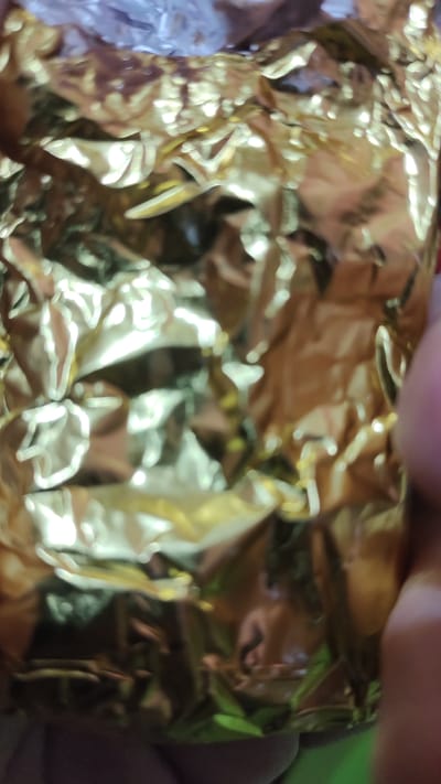 The image is a close-up, abstract shot of crumpled gold foil. The foil is highly reflective, creating distorted highlights and shadows that dominate the composition. There are no discernible objects, people, or environmental details clearly visible, only the textured surface of the foil. The lighting is harsh and appears to be coming from above, creating bright, blown-out areas and deep, dark shadows. The overall impression is one of texture and light play on a metallic surface. This image is not specific to Palangka Raya, Indonesia.