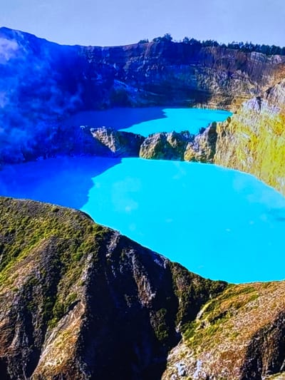 The image displays a dramatic natural scene with steep, rocky cliffs on either side of a bright blue body of water. The cliffs are textured, showing rough surfaces and variations in color, with patches of green vegetation clinging to them. The water is a vivid turquoise, reflecting the bright sky. In the foreground, the water appears deeper and darker blue, with swirling patterns suggesting movement or depth. The overall impression is one of a powerful natural landscape, possibly a fjord, canyon, or a deep lake. The lighting suggests it is daytime, with bright sunlight illuminating the scene. There is no visible text or people in the image.
