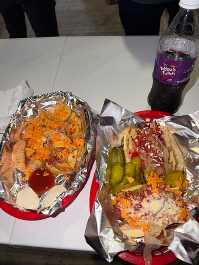 The image shows two red plastic baskets containing food, a bottle of soda, and part of a person. The food appears to be overloaded and messy, wrapped partially in foil. One basket contains what looks like a hot dog with toppings and several pickles. The other basket contains sliced potatoes with shredded orange and red toppings, along with small portions of ketchup and mayonnaise. A bottle of "Delaware Punch" soda is visible. Part of a person's leg is visible on the left side of the image, appearing to wear dark pants. The scene is set on a white table. Based on the type of food seen in this picture, this image has been taken in Tampico, Mexico.