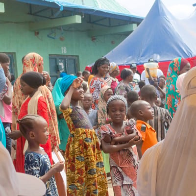 The image depicts a gathering of people, predominantly children, in Gudi, Nigeria. Many of them are wearing colorful, patterned clothing and head coverings. A blue tent is erected in the background, adjacent to a green building with the number "18" visible. Some adults are interspersed among the children, with at least one individual carrying a bucket. The overall scene suggests a community event or gathering.