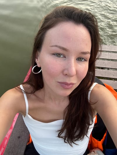 A woman with long, dark brown hair and green eyes is taking a selfie outdoors. She is wearing a white tank top and large silver hoop earrings. Her skin is fair, and she appears to be wearing light makeup. Behind her, there is calm water, and to her right, a wooden deck or pier. The woman is seated, with her arm visible in the foreground, and part of what appears to be an orange life vest is also visible. The image is taken from a slightly high angle. The setting suggests a waterside location, potentially a park or recreational area within the city of Ivanovo, Russia, given the context.