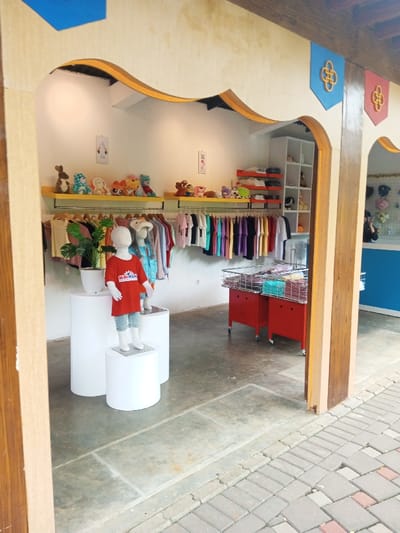The scene depicts the interior of a retail establishment, likely a children's store, viewed from an exterior paved area in Tân Kỳ Commune, Vietnam. The entrance structure features wooden archways and columns, adorned with blue and red banners each displaying a golden knot emblem. The store's interior has white walls, a grey concrete floor, and is illuminated by overhead recessed lighting.
In the foreground, three white child-sized mannequins stand on white cylindrical pedestals of varying heights. One mannequin, prominent in the center, wears a red short-sleeved t-shirt with a "MINI MAN" logo and light blue denim shorts. A green potted plant in a white pot is situated on a pedestal to the left. Along the back wall, two horizontal metal racks display numerous colorful t-shirts and other garments on hangers. Above these racks, a yellow shelf holds a diverse collection of stuffed animals and plush toys. To the right, two red mobile carts with metal basket tops contain folded clothing items. Additional white and orange shelving units, stocked with merchandise, are visible on the right side of the store. A blurry, indistinct human figure is faintly visible behind a blue counter in the far right background. The exterior ground consists of interlocked grey and reddish paving bricks.