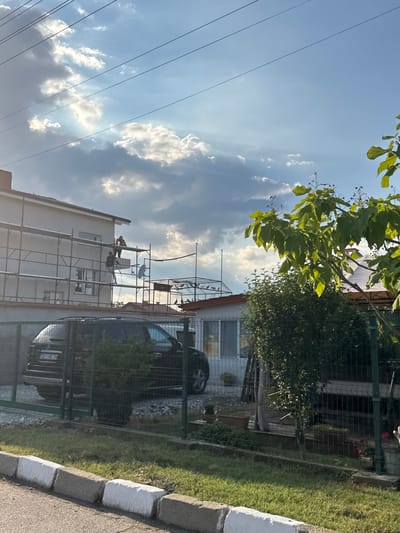 The image depicts a construction scene with workers on scaffolding, likely engaged in repairs or renovations on a building. Two individuals are visible on the scaffolding, one appearing to be active in the work while the other is positioned nearby. The building itself is light-colored and has several windows. Scaffolding surrounds a portion of the building, suggesting ongoing construction or maintenance.
Behind the building, a dark-colored car is partially visible through a metal fence. The car appears to have a license plate that reads "CB 1848 XK". Beyond the car, there is a grassy area and more fencing. In the foreground, a concrete curb with white stripes runs along an asphalt surface.
Above the scene, the sky is a prominent feature, filled with blue sky and scattered white and gray clouds. Sunlight streams through the clouds, creating illuminated patches. Green leaves and branches from a tree extend into the frame from the upper left corner, casting shadows and adding a natural element to the otherwise constructed environment. The overall lighting suggests it is daytime, possibly late afternoon given the angle of the sun and the shadows. The presence of scaffolding and workers indicates an active construction or repair event. The location is identified as Gelemenovo, Bulgaria.